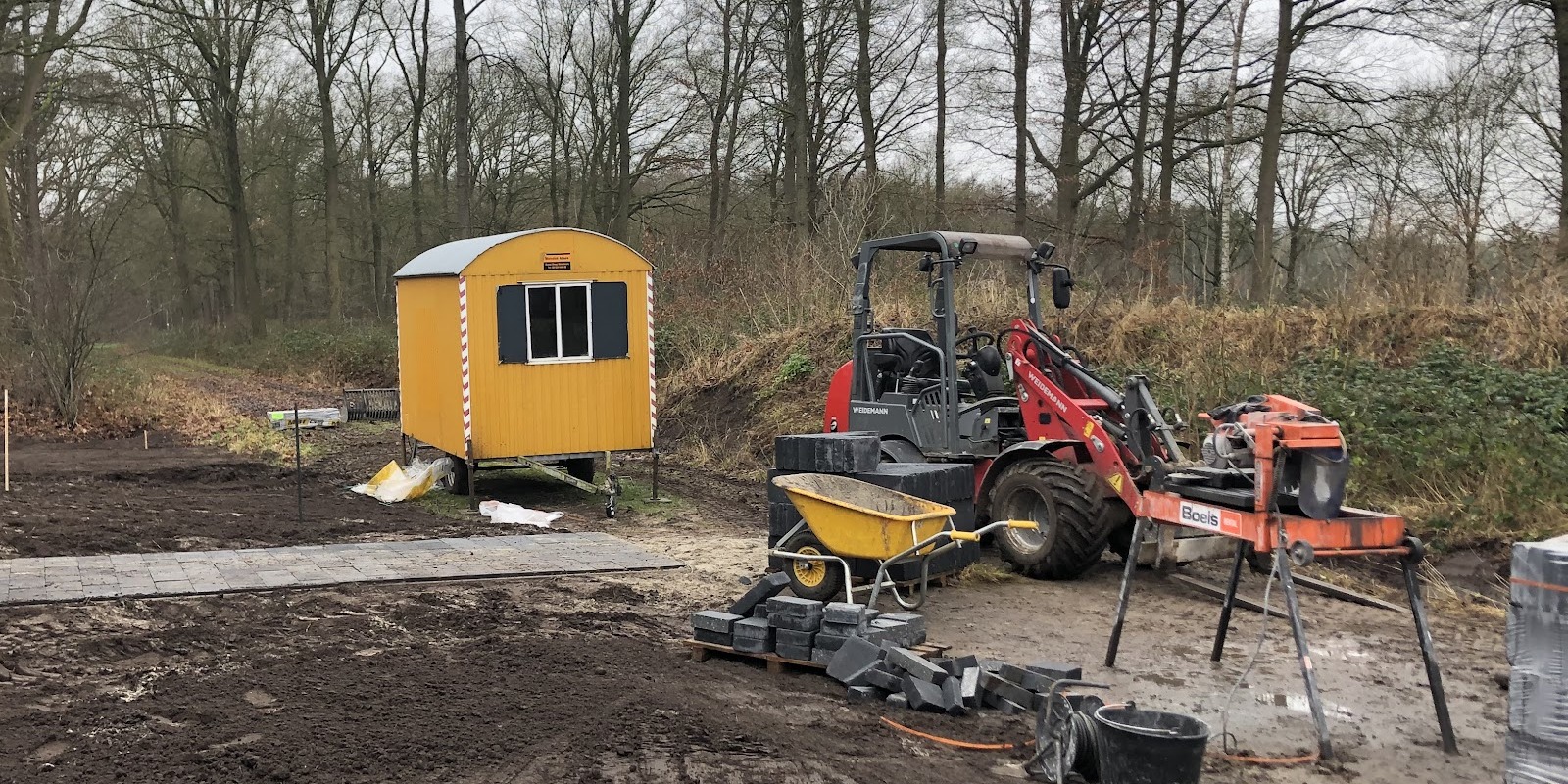 Bouwterrein met een minishovel en een gele schaftkeet, het grondwerk is bezig